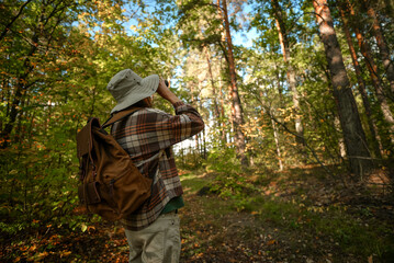 Rear view of a man in a hat standing in the woods and looking through binoculars