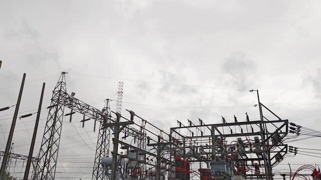 Low angle cinematic pan right of high voltage electrical substation switchyard with steel lattice structures and insulators against overcast sky.