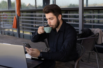 The man is drinking from a cup while looking at his phone, with a laptop open 