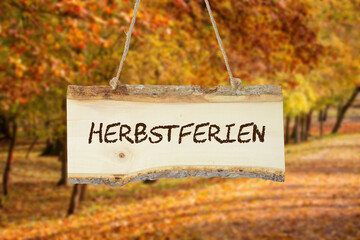 Holzschild mit der Aufschrift HERBSTFERIEN vor einem herbstlich gefärbten Laubwald