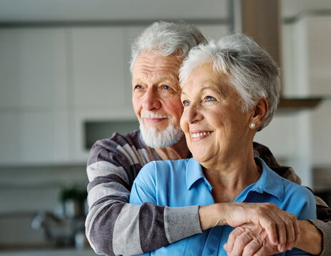 Portrait of a happy senior couple embracing hugging and bonding looking through the window at home - Powered by Adobe