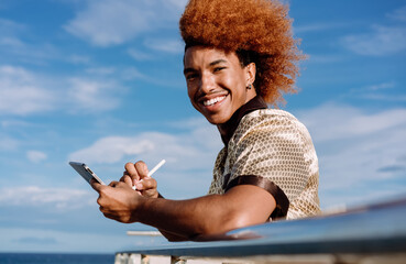 Smiling African American man with curly ginger afro outdoors, holding tablet and stylus, looking at camera, symbolizing confidence, technology adoption, and stylish mobile freelance culture.