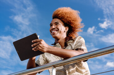 Smiling man with curly red afro holding tablet and stylus while outdoors, wearing patterned silk...