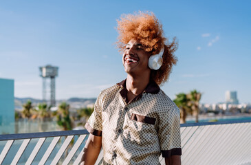 Smiling young man with curly afro listening to music on wireless headphones, looking up joyfully,...