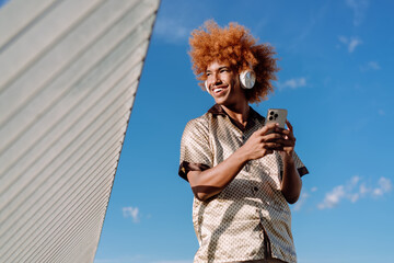 Joyful African American man with curly afro using wireless headphones, smartphone in hand, smiling...