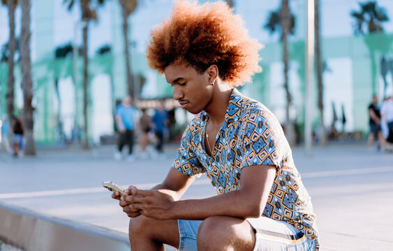 Fototapeta Young man with curly afro looking at smartphone with serious expression. Lifestyle photo showing focus, attention, and modern mobile interaction outdoors.