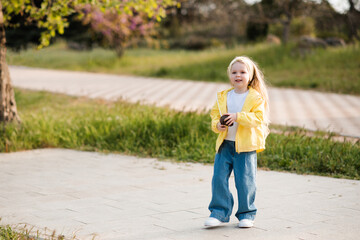 Fototapeta premium Funny smiling baby girl 2-3 year old wearing yellow autumn jacket and denim pants holding fir cone in park. Autumn season.