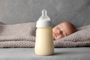 A full baby bottle sits in front of a sleeping infant wrapped in a gray blanket
