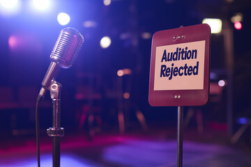 A microphone on a stand next to a sign that reads Audition Rejected in a dimly lit stage setting.