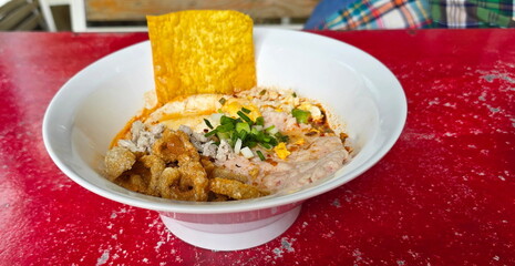 Spicy Tom Yum noodles served with large slices of minced pork, fried wontons, and pork crackling. Noodles in a white bowl are placed on a red table. The soup is made with chili paste and  lime juice.
