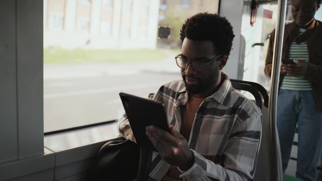 African American man wearing glasses holding tablet while riding bus. Passenger focused on screen learning or reading content. Another female standing in background checking phone. Public transport.