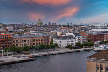 View of Frederik's Church with its green dome, a canal, historic and modern buildings, and a pink and blue sky in Copenhagen, Denmark.