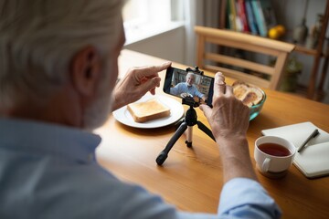 An elderly man uses a smartphone on a tripod to record himself while enjoying breakfast at a cozy kitchen table, creating a personal video.