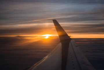 An airplane wing is visible from a window during sunset, with the sun casting a golden glow and a...