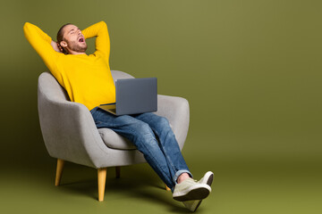 Young man in casual outfit relaxing on chair with a laptop in a stylish setting with a vibrant green khaki backdrop