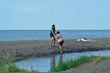 The daughter holds her motherâ€™s hand as they walk together from the shallow water stream onto the sandy beach, showing tenderness and care in a quiet tropical moment.