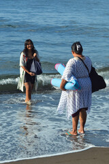 A teenage girl plays near gentle waves as her mother stands on the sandy shore carrying a bag and...