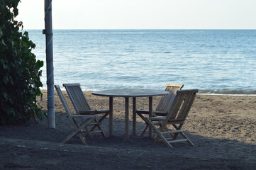 An outdoor beach cafe with wooden tables and chairs stands directly on sand facing calm sea, framed by greenery under bright clear daylight.