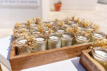 A tray filled with handmade candles in small jars is adorned with dried flowers. The scene is illuminated by bright sunlight, creating a warm and inviting atmosphere.