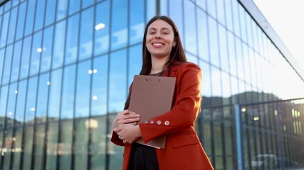 Portrait of a successful young professional woman holding a clipboard and smiling at the camera. She is standing outdoors with a contemporary glass office building behind her - Powered by Adobe