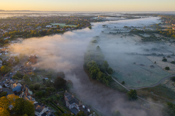 Aerial image of a Morning mist flows over the River Dee with nearby houses and fields in Chester at sunrise.