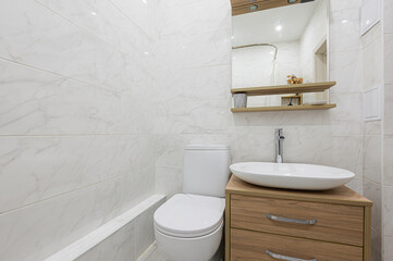 A modern bathroom interior featuring white marble tiles, a toilet, a sink on a wooden cabinet, and a mirror with shelves