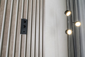 A modern interior view shows a wooden slat wall with a black outlet. A sleek floor lamp with three lights is positioned to the right of the wall against a neutral textured backdrop
