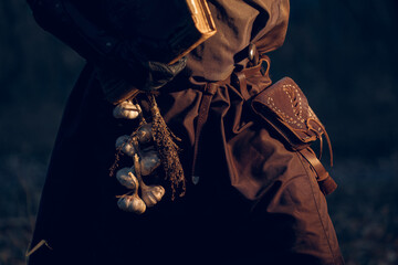 Mysterious figure cloaked in dark fabrics carries garlic, evoking a medieval or fantasy aesthetic.