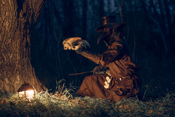 Mysterious plague doctor kneels beneath a tree, illuminated by lantern light and holding a skull in dark forest setting.