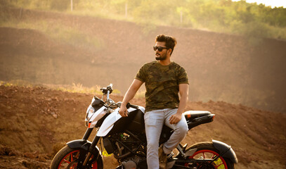 Young man in camouflage t-shirt sits on his motorcycle at sunset in rural India, creating a...