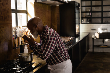 Senior African American man tasting soup over stainless steel pot on gas stove in modern kitchen