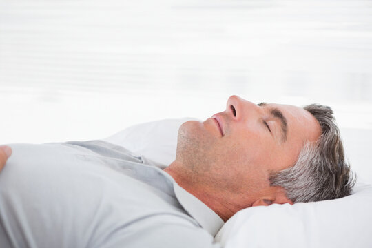 Middle-aged man resting on white bedding and pillow in bedroom wearing light grey button-up shirt