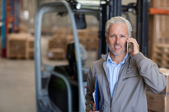 Senior manager speaking on phone, reviewing clipboard amid racks and parked forklift in warehouse