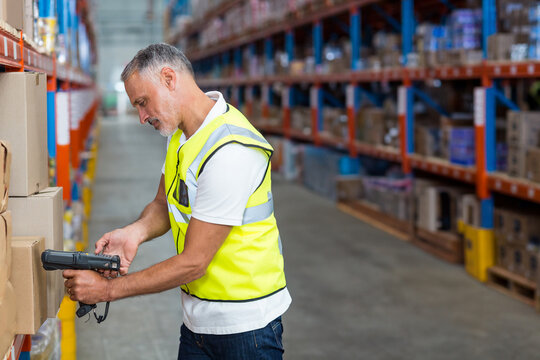 Handheld barcode scanner scanning cardboard box on tall metal shelving units in warehouse aisle