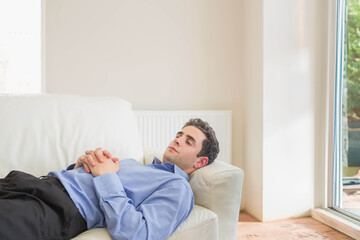 Man lying on light-colored leather sofa with cushion near radiator and large window in living room