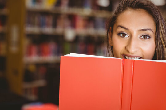 Female student holding up red hardcover book over face and peeking eyes at library shelves