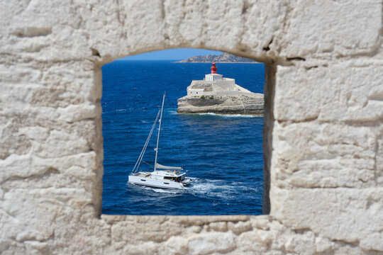 Fototapeta White sailboat passes near a lighthouse on a rocky island, seen through a stone window with bright blue sea.
