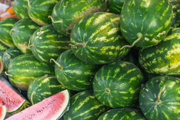 Fresh watermelons are piled high at an outdoor market, showcasing their vibrant green stripes against the warm summer sunlight. Nearby, sliced pieces offer a glimpse of juicy, red fruit
