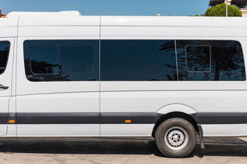 A large white passenger van is parked on a paved surface, surrounded by trees and buildings on a bright sunny day. The van features tinted windows and a sleek design © Creative_Bird
