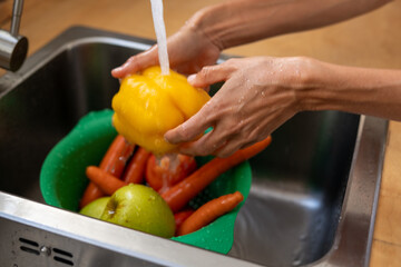 Hands washing fresh yellow bell pepper under running water in kitchen sink with carrots and apples in green colander