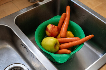 Fresh carrots, apple, nectarine, and yellow bell pepper in green colander inside kitchen sink with water drops after washing.
