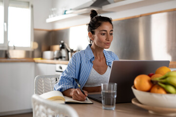 Young woman working on laptop in kitchen while writing notes in notebook, wearing striped pajamas with bowl of fresh fruit and glass of water on wooden table