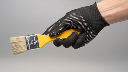 Hand in work glove holding yellow paintbrush on gray background