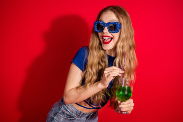 Smiling woman with stylish sunglasses enjoying a drink against a vibrant red background