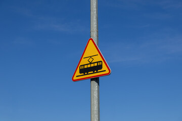 Yellow triangular tram warning sign on a metal pole against clear blue sky, indicating public transportation crossing and essential road safety