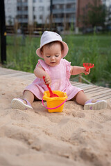adorable baby in pink dress enjoys playful moment with bucket and spade at urban playground sandpit, surrounded by vibrant greenery and residential buildings, happiness, discovery, interaction