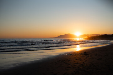 Scenic beach sunset with glowing sun over calm ocean waves, reflecting golden light on wet sand and distant mountains.