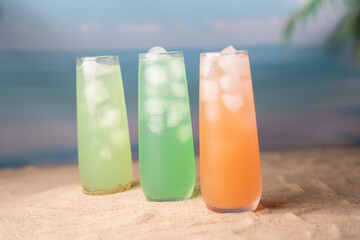three refreshing ice-filled glasses with colorful beverages on sandy beach, evoking a tropical ambiance under a soft blue sky with hints of blurred palm fronds, idyllic, backdrop, peaceful, horizon