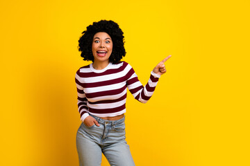 Young woman with curly hair in casual outfit joyfully pointing on yellow background showcasing positive emotion