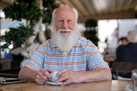 Old man with a long white beard sitting in a café, smiling warmly while holding a cup of coffee at a wooden table, surrounded by a cozy atmosphere